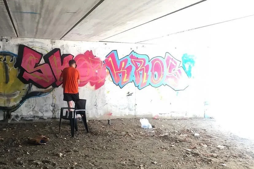 Artist standing on a chair painting pink and blue graffiti lettering across an unfinished interior wall