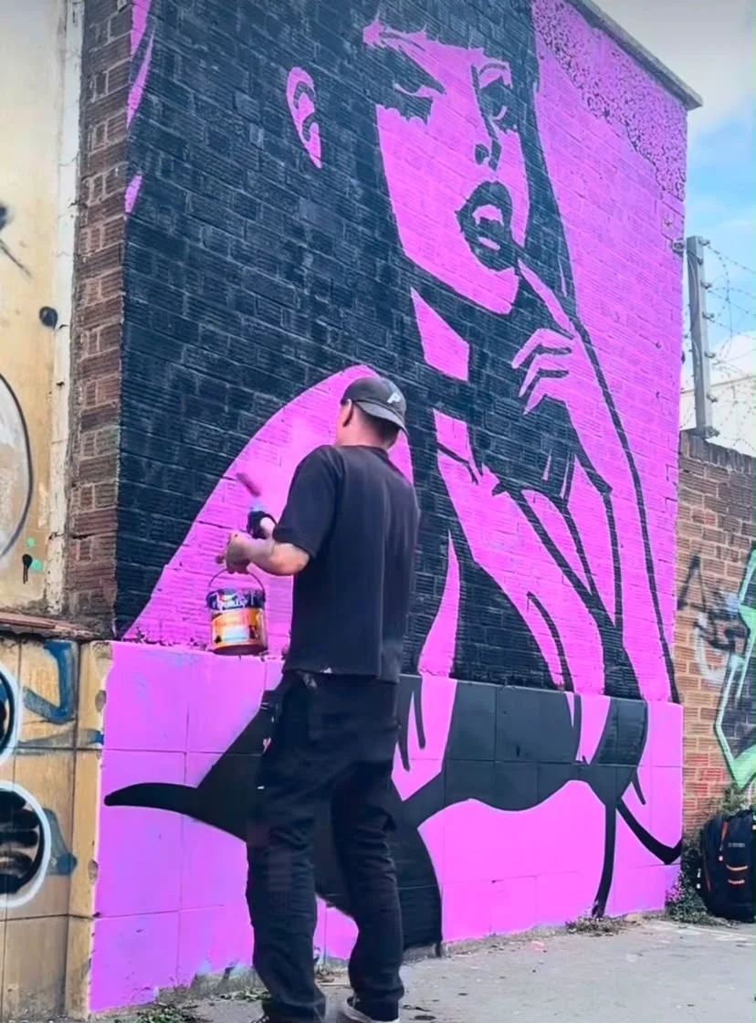 Painter standing in front of a large pink and black portrait mural in progress on a brick wall